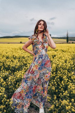 Güzel, siyah saçlı, zarif elbiseli, çiçek açan kolza tohumu tarlasında poz veren güzel bir kadının moda fotoğrafı.