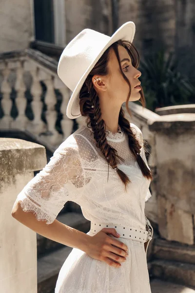 エレガントな白いドレスと階段でポーズをとる帽子の暗い髪の美しい女性のファッション屋外写真 — ストック写真