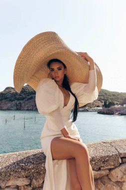 Güzel, siyah saçlı, beyaz elbiseli ve şapkalı güzel bir kadının moda fotoğrafı. Deniz manzaralı güzel bir poz veriyor.