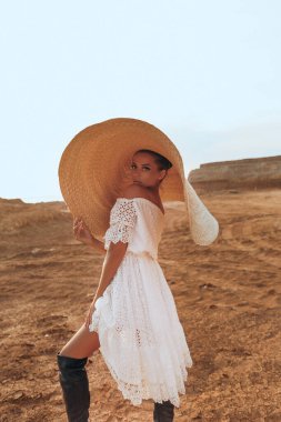 Güzel, sarışın, zarif elbiseli ve çölde poz veren büyük hasır şapkalı seksi bir kadının moda fotoğrafı. 