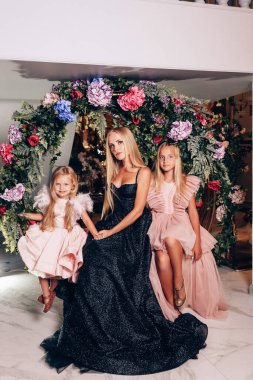 Annelerinin ve kızlarının şık elbiselerle süslü stüdyoda çiçek kemeriyle poz verdikleri güzel bir aile fotoğrafı.