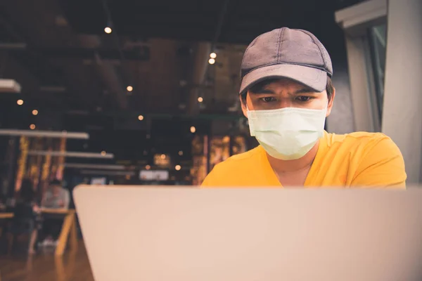 A man in yellow shirt and medical mask using laptop and smartphone while sitting in coffeeshop