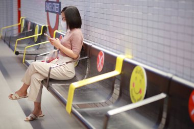 Tren istasyonunda oturan ve elinde cep telefonu olan Asyalı bir kadın maske takıyor ve Corona virüsü kapma riski olan COVID-19 enfeksiyonu yüzünden maske takıyor. Yeni normal kavramı.