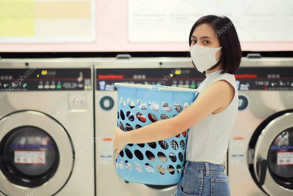 woman wearing a mask in quarantine for corona virus COVID-19 spreading outbreak. Doing laundry at laundromat shop during. New normal lifestyle social distancing for infection risk.