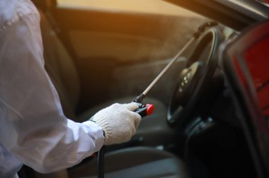 PPE seti giyen virolog arabada dezenfektan spreyi sıkar, Covid-19 virüsünün enfeksiyonunu önler. Mikrop ve bakterilerin kirlenmesi, sık sık dokunulan temiz yüzeyleri siler..