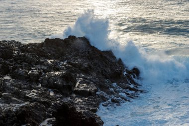 Güçlü dalga deniz kenarı lav kayaları günbatımında karşılamaktadır.
