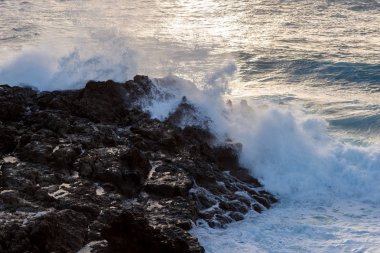 Güçlü dalga deniz kenarı lav kayaları günbatımında karşılamaktadır.