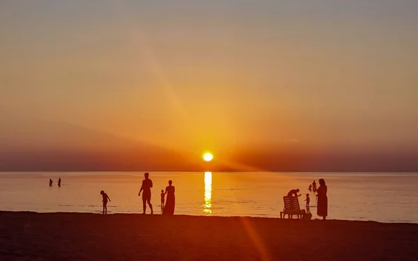 Akşam deniz kenarında, gün batımında, kıyı şeridine yakın karanlık insan siluetleriyle. Sahne, parlayan gökyüzü ve yansıtıcı suyla huzurlu ama dramatik bir atmosfer yakalıyor..