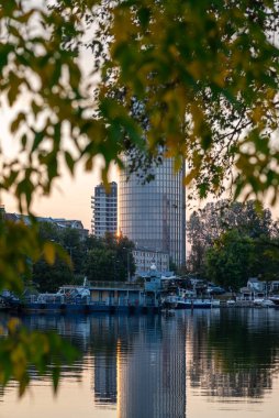 City view featuring a modern high-rise building reflected in calm canal waters, with several moored yachts in the foreground. Contemporary urban waterfront scene captured on a clear day.
