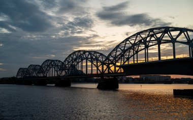 A railway bridge spanning over a river at sunset, with warm golden light reflecting on the water. The scene captures the tranquility of the landscape combined with the strength and structure of industrial architecture.