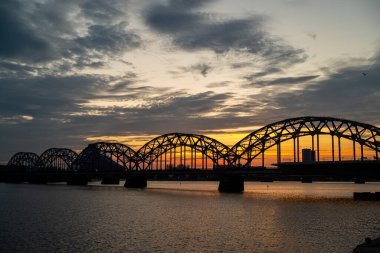 A railway bridge spanning over a river at sunset, with warm golden light reflecting on the water. The scene captures the tranquility of the landscape combined with the strength and structure of industrial architecture.