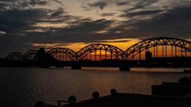 Extremely beautiful and peaceful scene of the Iron Bridge (Dzelzcea tilts) over the Daugava River at sunset, with its round iron arches standing out against the vibrant orange and blue evening sky.