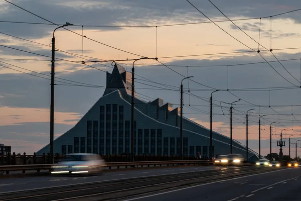 Real photograph of the National Library of Latvia viewed from the Stone Bridge at sunset, capturing warm evening light and architectural details.