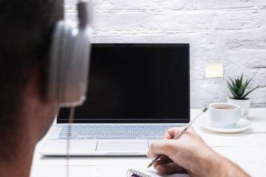 Kulaklıklı bir adam dizüstü bilgisayara bakarken not alıyor ve beyaz arka planda kahve içiyor..