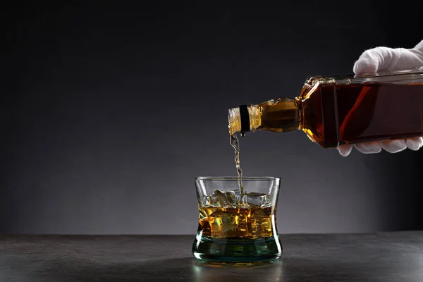 White gloved hand pouring whisky into a glass with ice on dark background, with copy space.