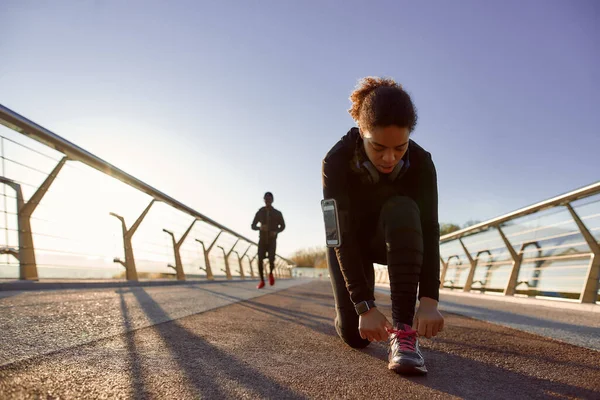 Ετοιμάζομαι για τζόγκινγκ. Αφρικανική αθλητική γυναίκα με μαύρα αθλητικά να δένει κορδόνια πριν τρέξει με φίλο ή φίλο στη γέφυρα το πρωί — Φωτογραφία Αρχείου