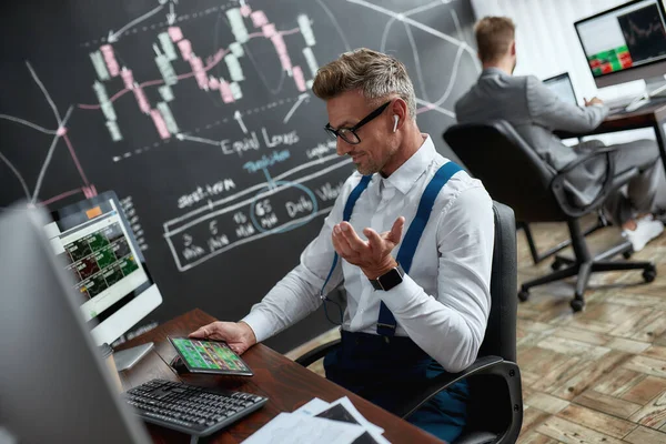 Het vinden van meer mogelijkheden. Stijlvolle zakenman, handelaar met behulp van zijn tablet pc, terwijl zitten aan het bureau voor computer monitor. Zijn collega werkt op de achtergrond — Stockfoto