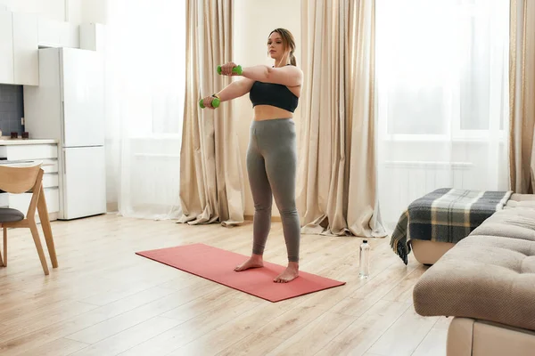 Celebrando la salud. Tiro de cuerpo entero de mujer joven con curvas en ropa deportiva haciendo ejercicio usando pesas en una esterilla de yoga en casa — Foto de Stock
