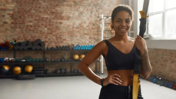 Sii una persona in forma. Donna sportiva che sorride alla macchina fotografica, tiene le cinghie fitness TRX mentre fa allenamento in palestra industriale — Foto Stock