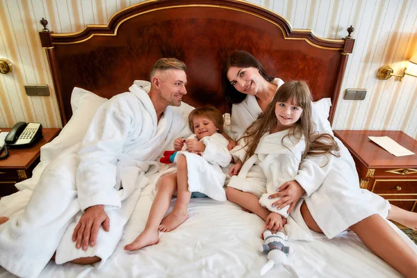 Familia feliz. Los padres caucásicos y dos niños en batas blancas sonriendo mientras yacen en la cama en la lujosa habitación de hotel. Familia, viajes, resort, concepto de vacaciones — Foto de Stock