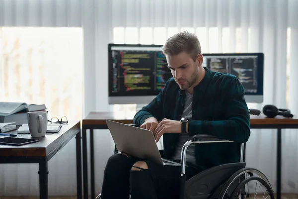 Een drukke dag. Portret van een jonge geconcentreerde mannelijke webontwikkelaar in een rolstoel die programmacode schrijft op zijn laptop op zijn werkplek. Meerdere computerschermen met programmacode — Stockfoto