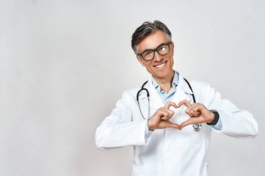 Üniforma ve gözlük takan, boynunda steteskop olan mutlu, olgun bir erkek doktor. Kalp işaretini gösteriyor ve gülümsüyor.