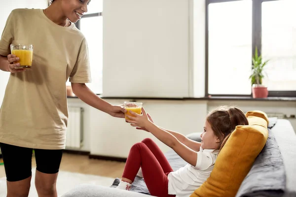 Estar sano. Mujer afroamericana niñera entretenido caucásico linda niña. El chico está sentado en el sofá mientras la niñera le da un vaso de jugo. —  Fotos de Stock