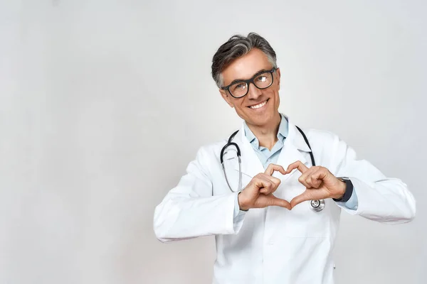 Ευτυχισμένος ώριμος άνδρας γιατρός φορώντας στολή και γυαλιά, με στηθοσκόπιο γύρω από το λαιμό που δείχνει το σημάδι της καρδιάς και χαμογελά, στέκεται πάνω σε γκρι φόντο — Φωτογραφία Αρχείου