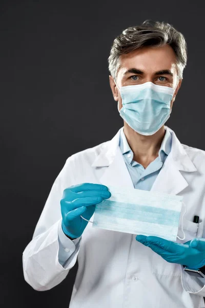 Kendini enfeksiyonlara karşı koru. Tıbbi üniformalı ve mavi steril eldivenli olgun bir erkek doktor siyah arka planda dururken kameraya tıbbi maske takıyor. — Stok fotoğraf