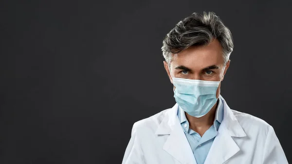 Koruyucu ekipman. Tıbbi üniforma giyen ve koruyucu maske takan kendine güvenen olgun bir erkek doktor siyah arka planda dururken kameraya bakıyor. — Stok fotoğraf