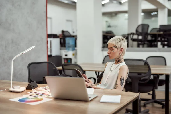 Making work schedule. Young and attractive tattooed business woman with short haircut using digital tablet while sitting at her workplace in the office