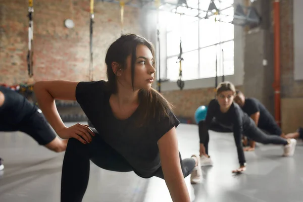 Recebe o corpo que mereces. Jovem mulher de sportswear preto olhar focado durante o exercício na academia industrial. Conceito de formação em grupo — Fotografia de Stock