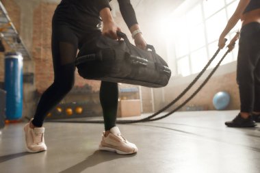 Defeat your weakness. Cropped shot of sportive woman lifting a heavy sandbag while having workout at industrial gym. Group training, teamwork concept clipart