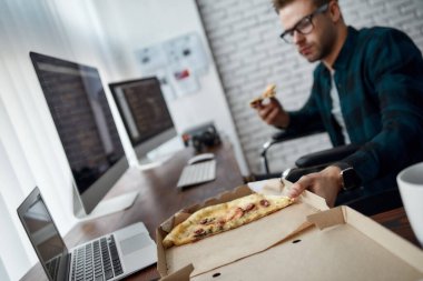 Mola zamanı. Modern ofisteki iş yerinde otururken tekerlekli sandalyede pizza yiyen genç bir web geliştiricisi. Engelli erkek programcı öğle yemeği yiyor.
