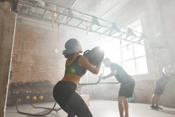 Tutkunla yaşa. Sportif bir kadın endüstriyel spor salonunda çalışırken kum torbasını kaldırıyor. Grup eğitimi, takım çalışması konsepti — Stok fotoğraf