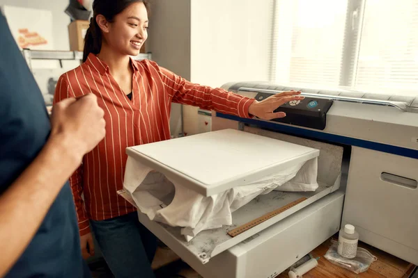 Facilita el trabajo. Trabajadores jóvenes, hombres y mujeres están preparando la camiseta para imprimir en la máquina de serigrafía en el lugar de trabajo — Foto de Stock