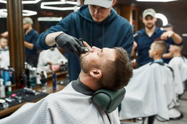 Bıyık Düzeltme. Berber, müşterisinin bıyığını elektrikli matkapla kesiyor. Berber dükkanını ziyaret eden genç bir adam. Yan görünüm