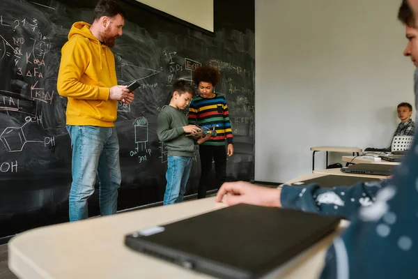 卓越中心。年轻的男性科学老师站在黑板旁边，拿着平板电脑，看着他的学生展示他们自己的机器人车 — 图库照片