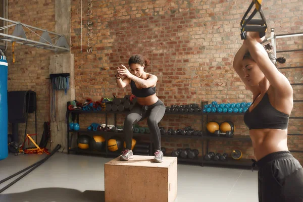 Essere in forma è una necessità. Donna sportiva che salta usando plyo box mentre fa allenamento in palestra industriale. Formazione di gruppo, concetto di lavoro di squadra — Foto Stock