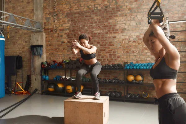 Estar em forma é uma necessidade. Mulher esportiva pulando usando caixa de plyo ao ter treino no ginásio industrial. Formação em grupo, conceito de trabalho em equipa — Fotografia de Stock