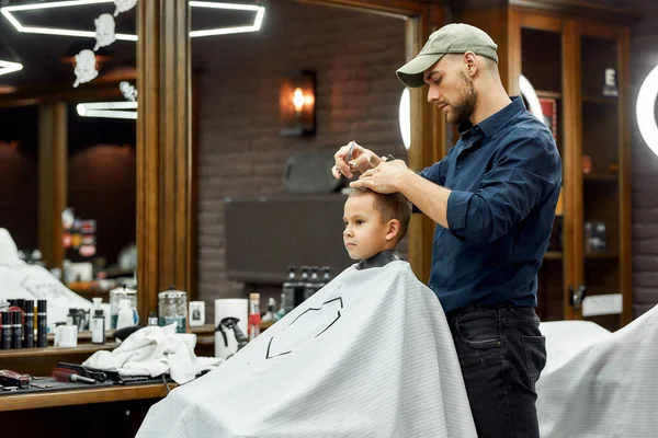 Stylish haircut for kids. Young male barber making a haircut for cute little boy in the modern barbershop