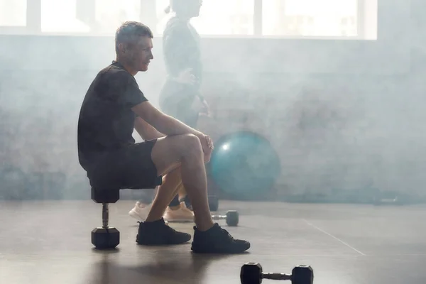 Blijf er aan werken. Moe atletische man zitten op een halter na het hebben van training in de industriële sportschool. Groepstraining, concept van gezonde levensstijl — Stockfoto