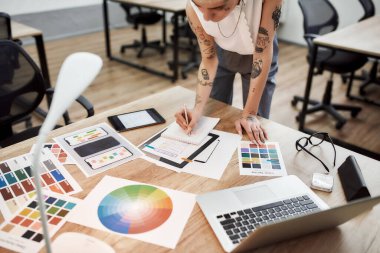 Tasarımcı işyeri. Ofis masasının yanında duran dövmeli kadın tasarımcının çizimleri. Renk örnekleriyle çalışmak