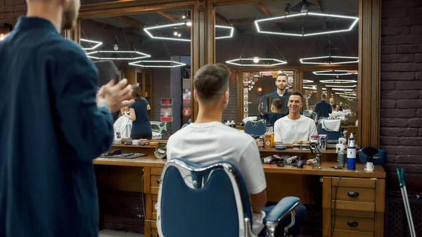 Cool Mens Penteado. Jovem feliz sentado em poltrona na barbearia e sorrindo enquanto barbeiro segurando um espelho e mostrando-lhe o resultado do trabalho. Novo corte de cabelo — Fotografia de Stock