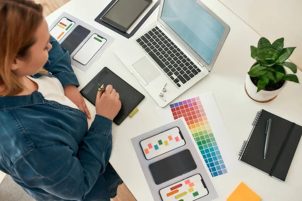 Vista dall'alto di giovane creativo designer ux ui femminile disegno su tavoletta grafica, lavorando da casa, seduto sul suo posto di lavoro. Scrivania con computer portatile, tamponi a colori, tavolozze e schizzi — Foto Stock