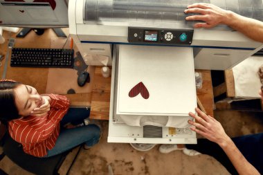 Kişiselleştirin. İş yerindeki ipek ekran baskı makinesinde tişört basılmasının sonucuna bakan genç işçiler, erkek ve kadınların üst görünümü