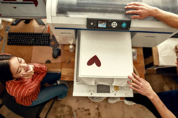 Hazlo personalizado. Vista superior de los trabajadores jóvenes, hombre y mujer mirando el resultado de la impresión de la camiseta en la máquina de serigrafía en el lugar de trabajo — Foto de Stock