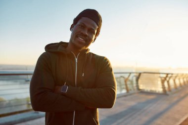 Koşmaya hazır. Siyah spor kıyafetli mutlu bir Afrikalı erkek koşucu kameraya gülümsüyor, sabah köprüde duruyor.