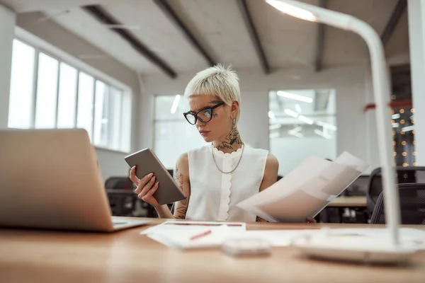 Making business easier. Young and beautiful tattooed business woman wearing glasses working with digital tablet while sitting at her workplace in the office