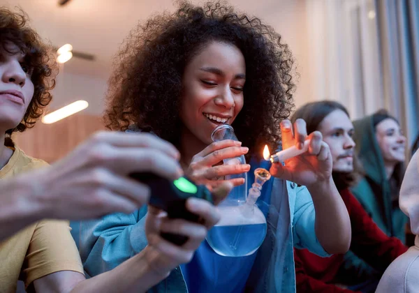 Para um bom humor. Jovem afro-americana acendendo maconha no cachimbo de vidro, relaxando com os amigos no sofá em casa. Jovens jogando videogames e fumando maconha — Fotografia de Stock